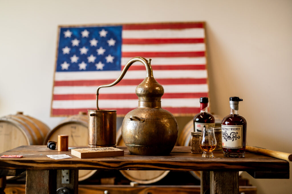 Copper still next to American flag and barrels for distilled spirits.